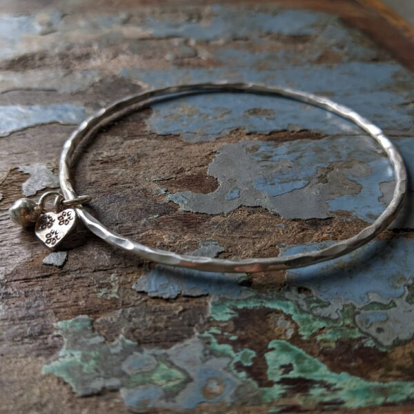 B016 Belle Bangle. Complete and continuous round bangle with a hammered and textured finish. Completed with a small round charm and an additional hallmarked heart charm.